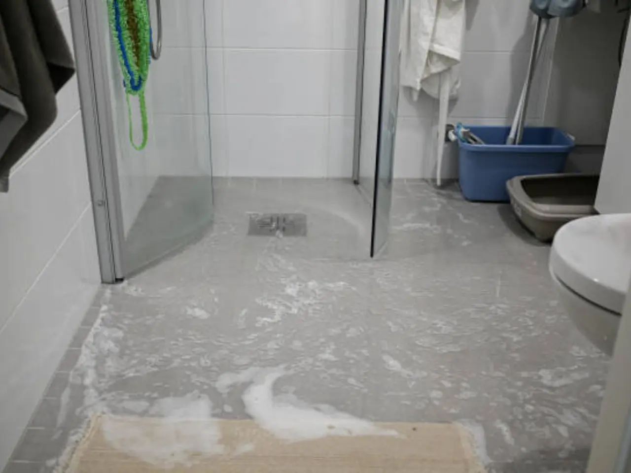 A bathroom with soapy water covering the grey tile floor, likely from an overflow. A shower, a toilet, a beige mat, and cleaning tools are visible in the room.