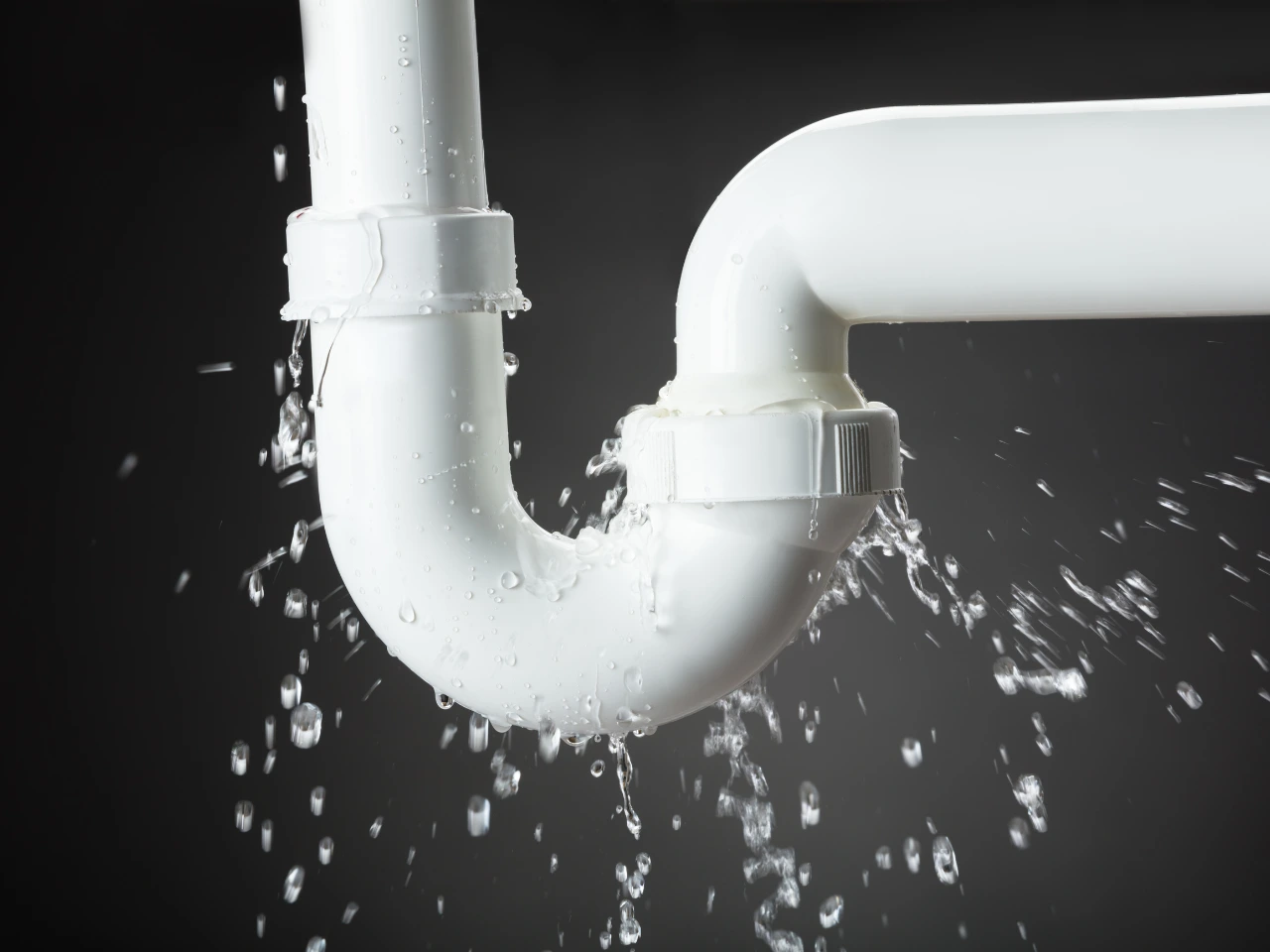 White plastic pipe with a U-bend leaking water at a joint, with water droplets spraying out, set against a dark background.