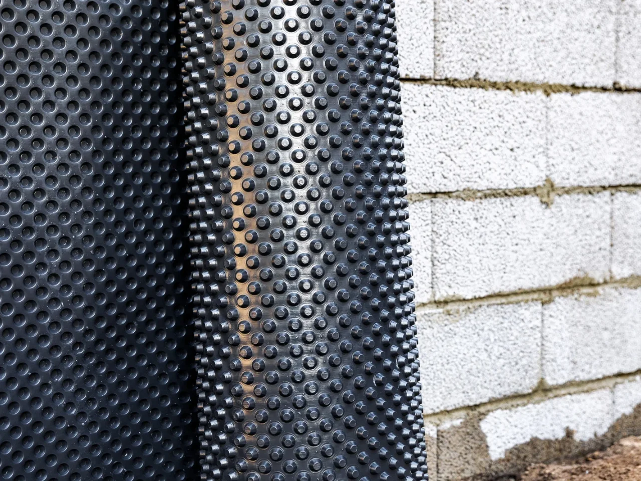 A close-up of a black dimpled waterproof membrane leaning against a white concrete cinder block wall, typically used for foundation protection in construction.