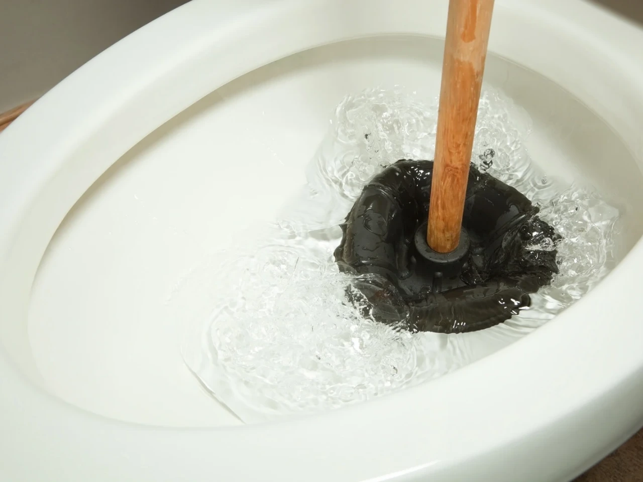 A close-up of a toilet being plunged with a black rubber plunger, causing water to swirl and splash inside the bowl.