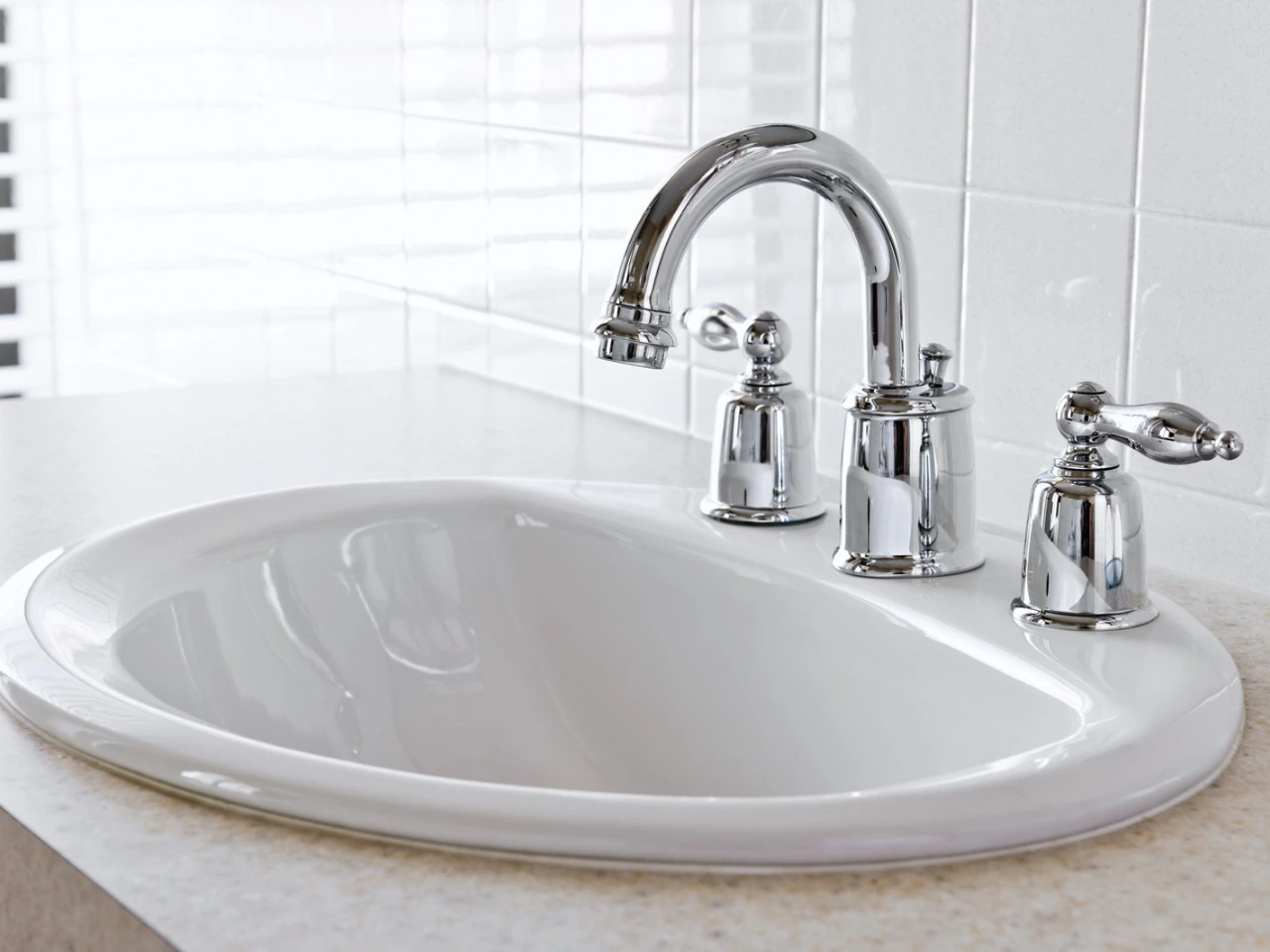 A clean, white bathroom sink with a shiny silver faucet and two handles sits on a light-colored countertop against a white tiled wall.