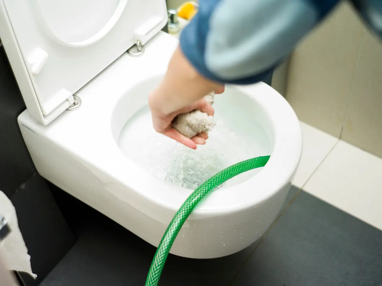 A person’s hand holding a cloth, cleaning the inside of a white toilet bowl with water flowing from a green hose. The toilet lid is open and a roll of toilet paper is visible on the left.