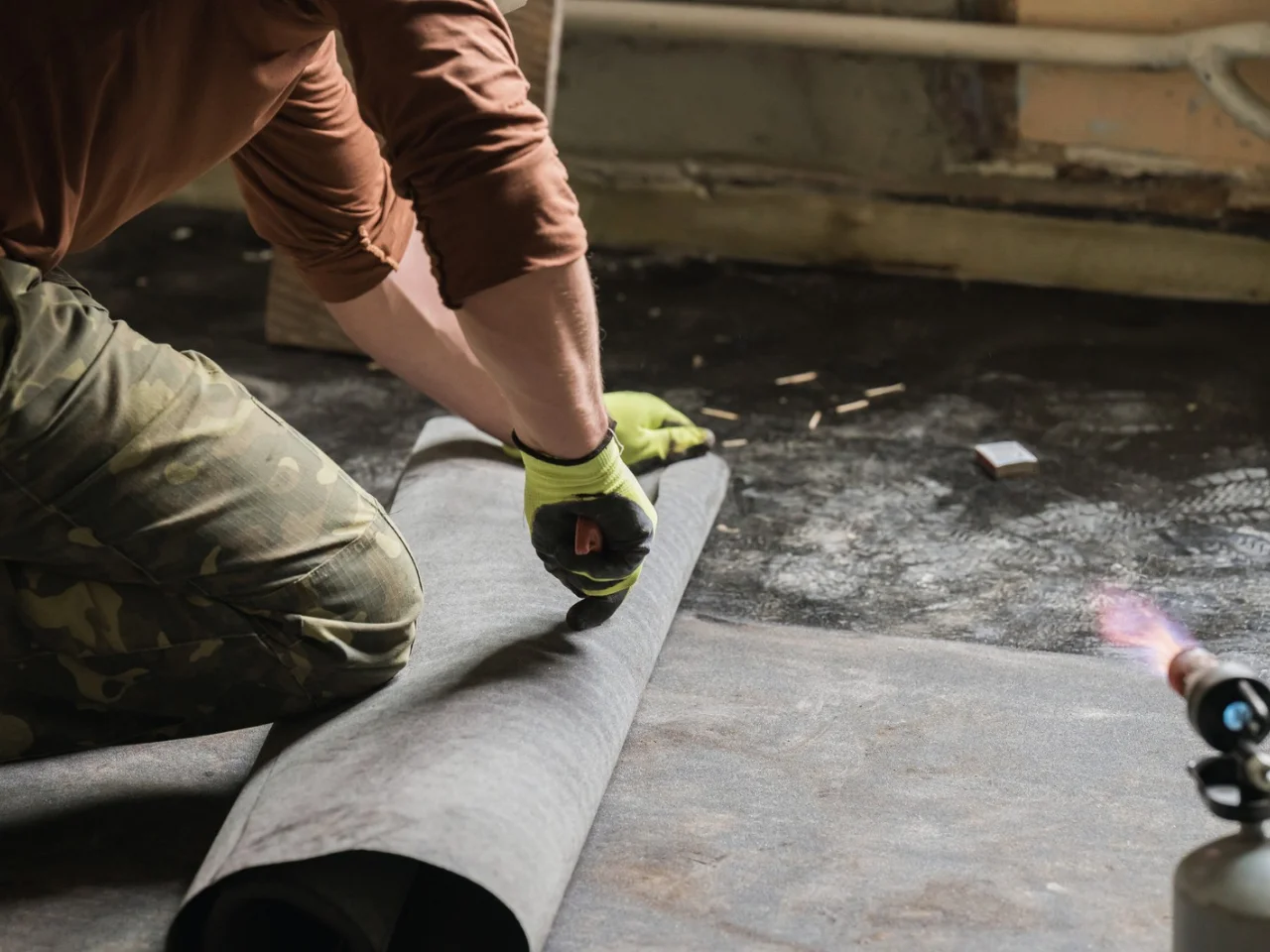 A person wearing camouflage pants and gloves kneels on the ground, preparing to cut or lay down a roll of roofing material or felt, with a torch for heating visible nearby on a dark surface.