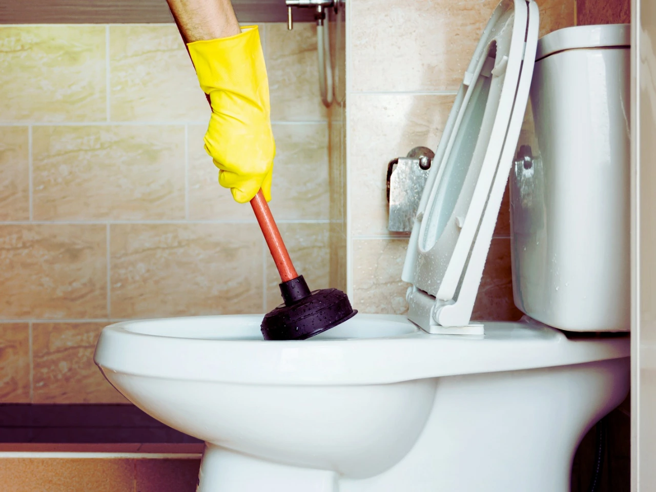 A person wearing a yellow rubber glove uses a plunger to unclog a white toilet in a tiled bathroom. The toilet lid is open.