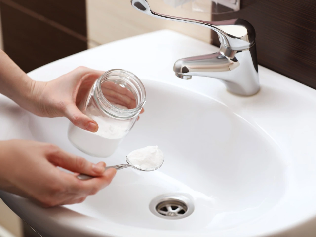 A person holds a small glass jar of white powder and scoops some with a spoon over a white bathroom sink with a silver faucet.