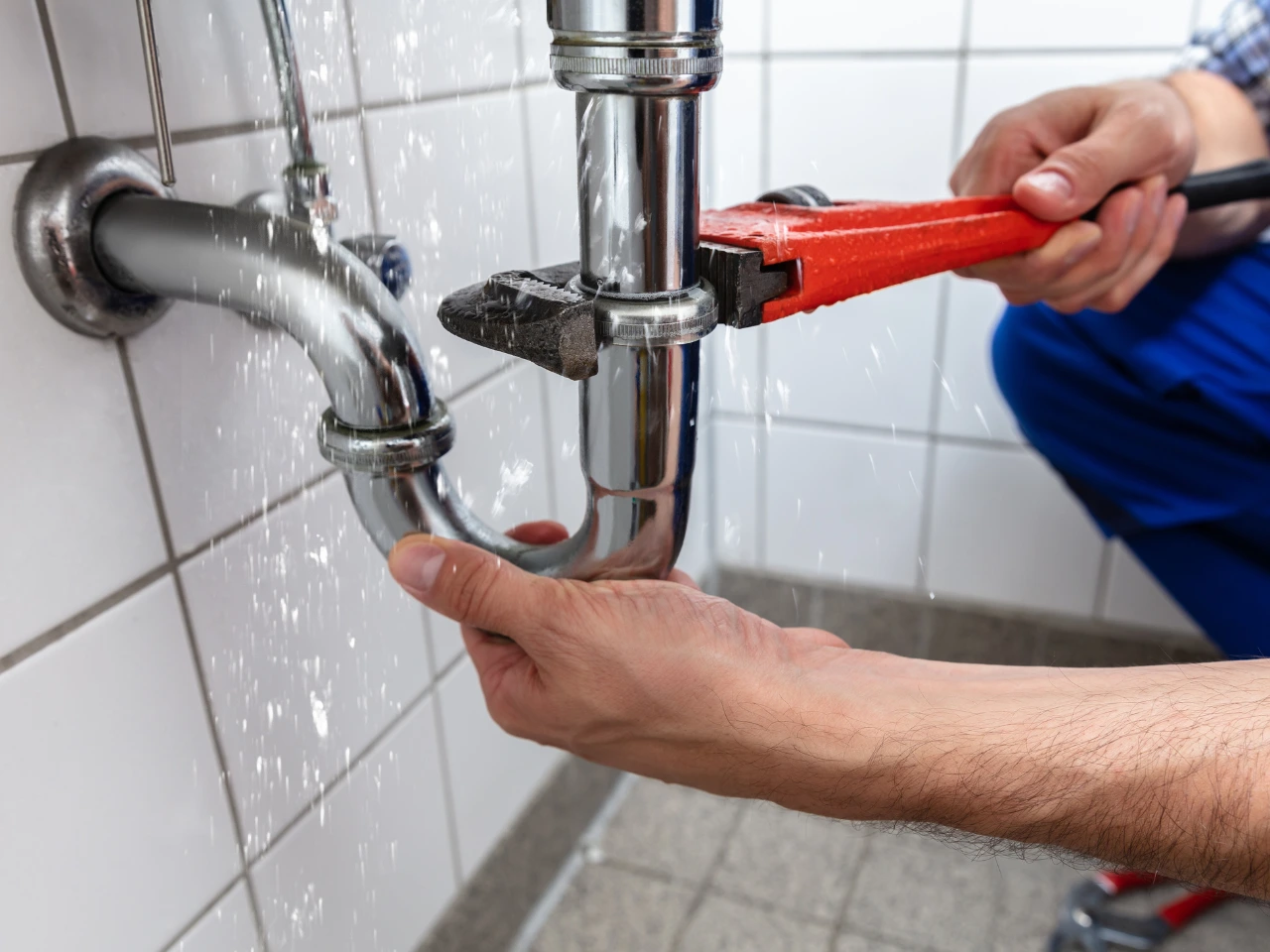 A person uses a red pipe wrench to fix a leaking chrome pipe under a sink, with water spraying out, in a tiled room.