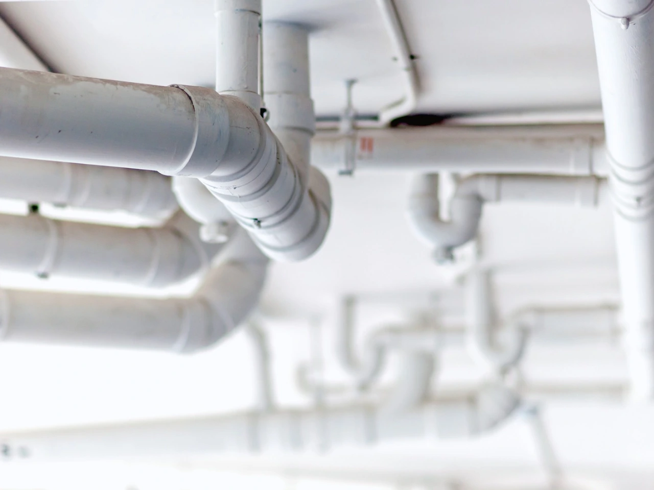 White plumbing pipes run across a white ceiling, intersecting and bending at various angles, creating an intricate network of tubes and fittings. The image focuses on the pipes, with a softly blurred background.