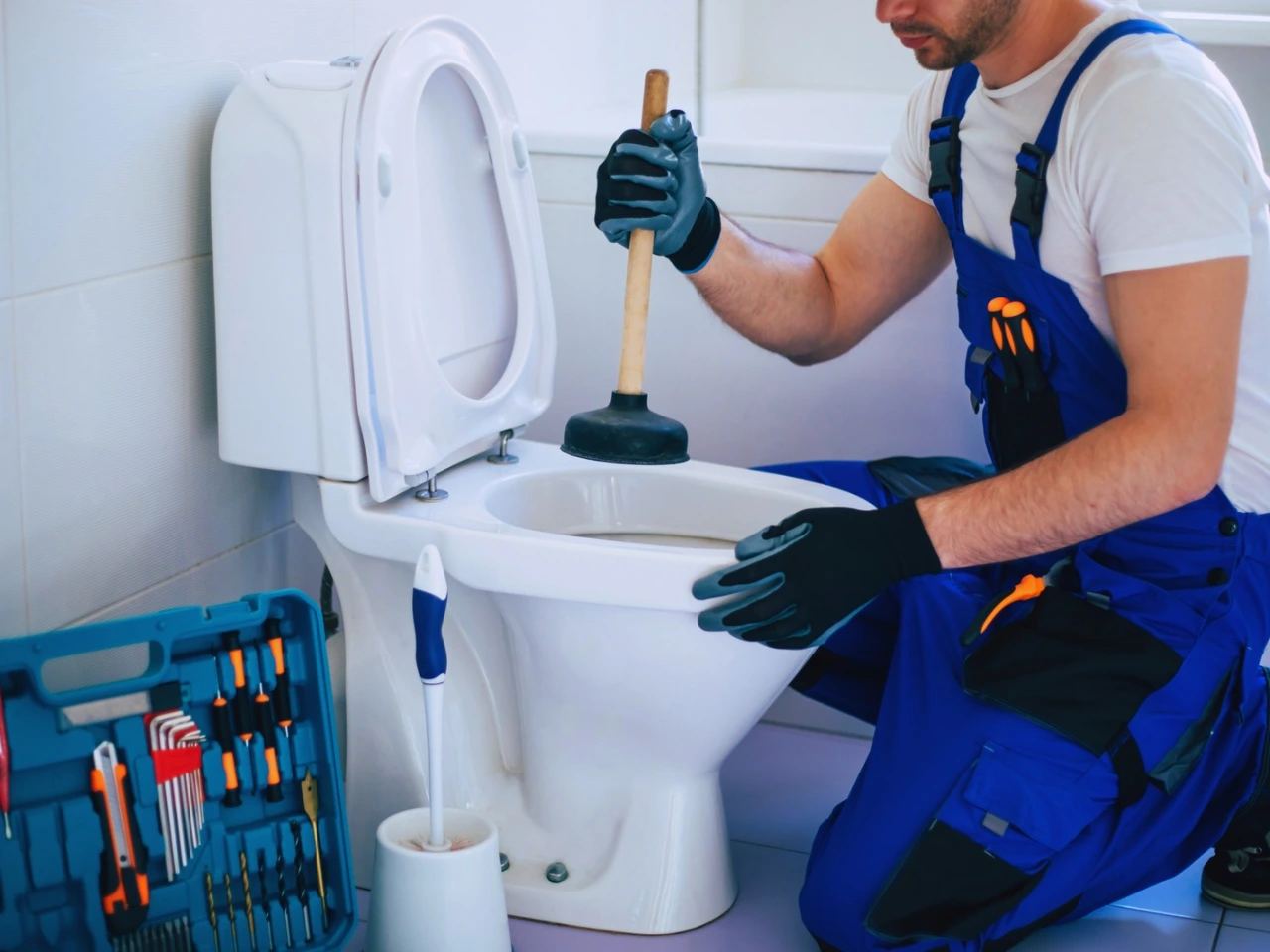 A plumber wearing blue overalls and black gloves uses a plunger on a toilet. A toolbox with various tools and a toilet brush are on the floor nearby.