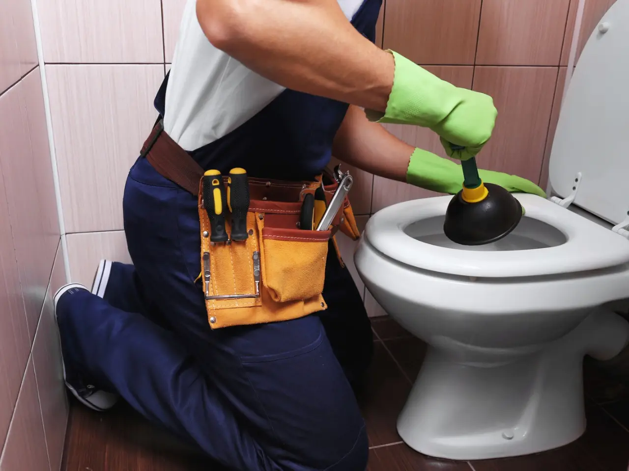 A person wearing green gloves and a tool belt kneels on the floor, using a plunger to unclog a toilet in a tiled bathroom.