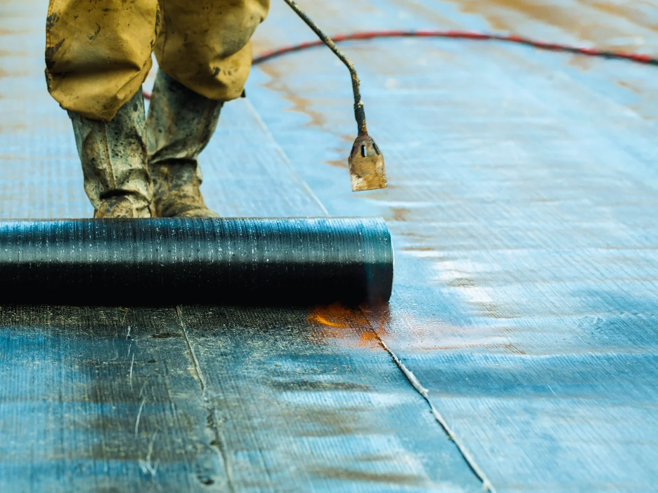 A worker in boots unrolls a sheet of waterproofing material onto a flat surface, using a gas torch to apply or seal it, likely as part of a roofing or construction project.