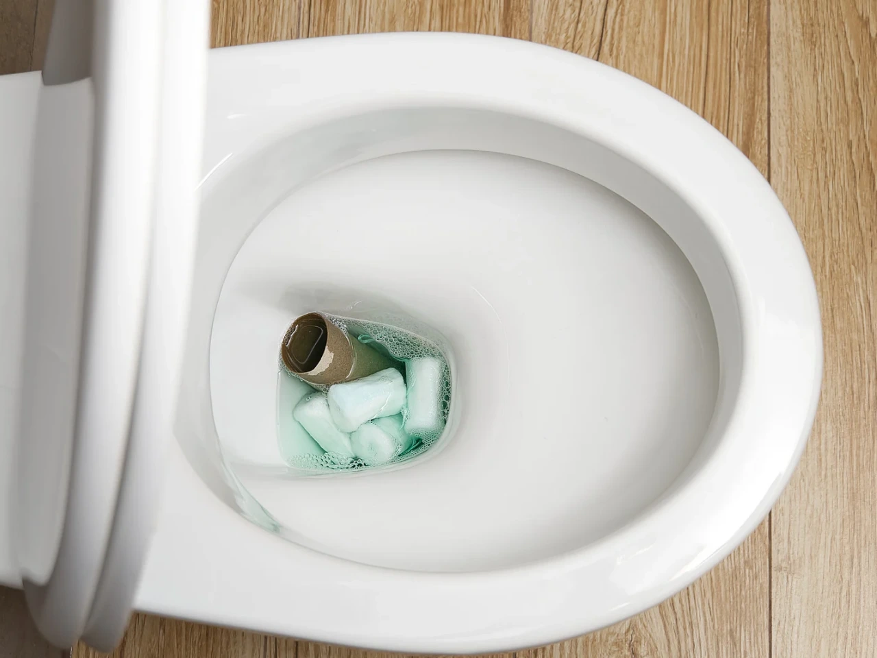 A toilet bowl containing a used cardboard toilet paper roll and several green cleaning tablets, with bubbles forming around them. The toilet seat is up, and the floor is wooden.