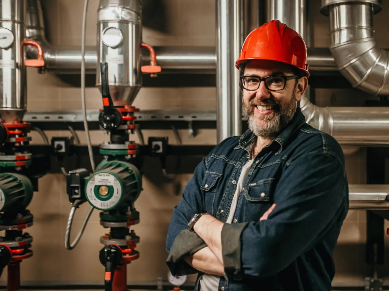 A man wearing a red hard hat, glasses, and a denim shirt stands smiling with arms crossed in front of industrial pipes and valves in a mechanical or boiler room.