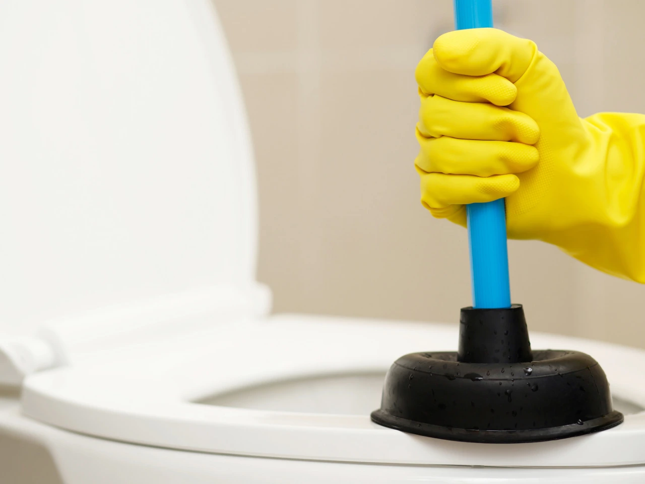 A person wearing yellow rubber gloves is holding a blue-handled plunger inside a white toilet bowl with the lid raised.