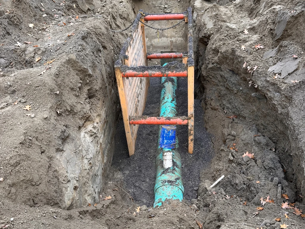 A green underground pipe is exposed in a trench during drain repair Etobicoke, with a wooden and metal safety barrier in place. Soil surrounds the trench, and scattered fallen leaves rest on the ground.