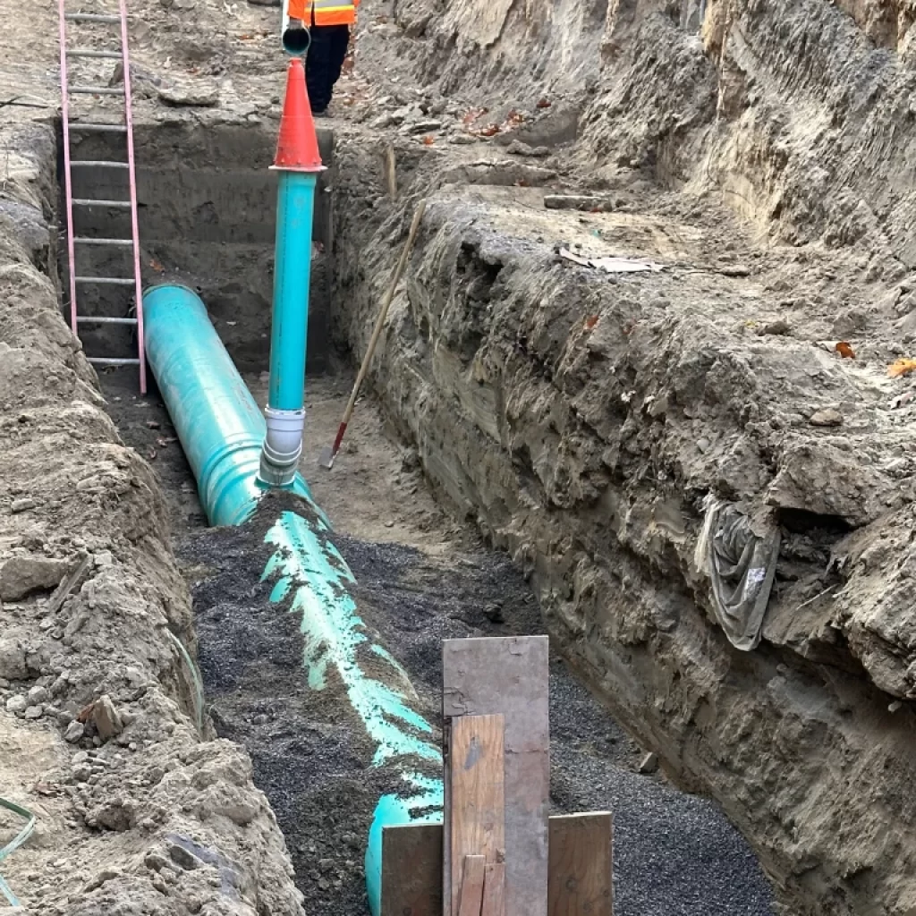 A construction worker stands in a deep trench next to large green pipes being installed underground, part of drain repair and plumbing in Toronto. A red and green vertical pipe rises above the trench, and a ladder leans against the dirt wall.