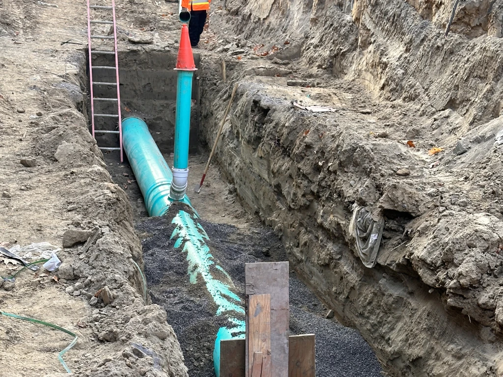 A construction worker stands in a deep trench next to large green pipes being installed underground, part of drain repair and plumbing in Toronto. A red and green vertical pipe rises above the trench, and a ladder leans against the dirt wall.