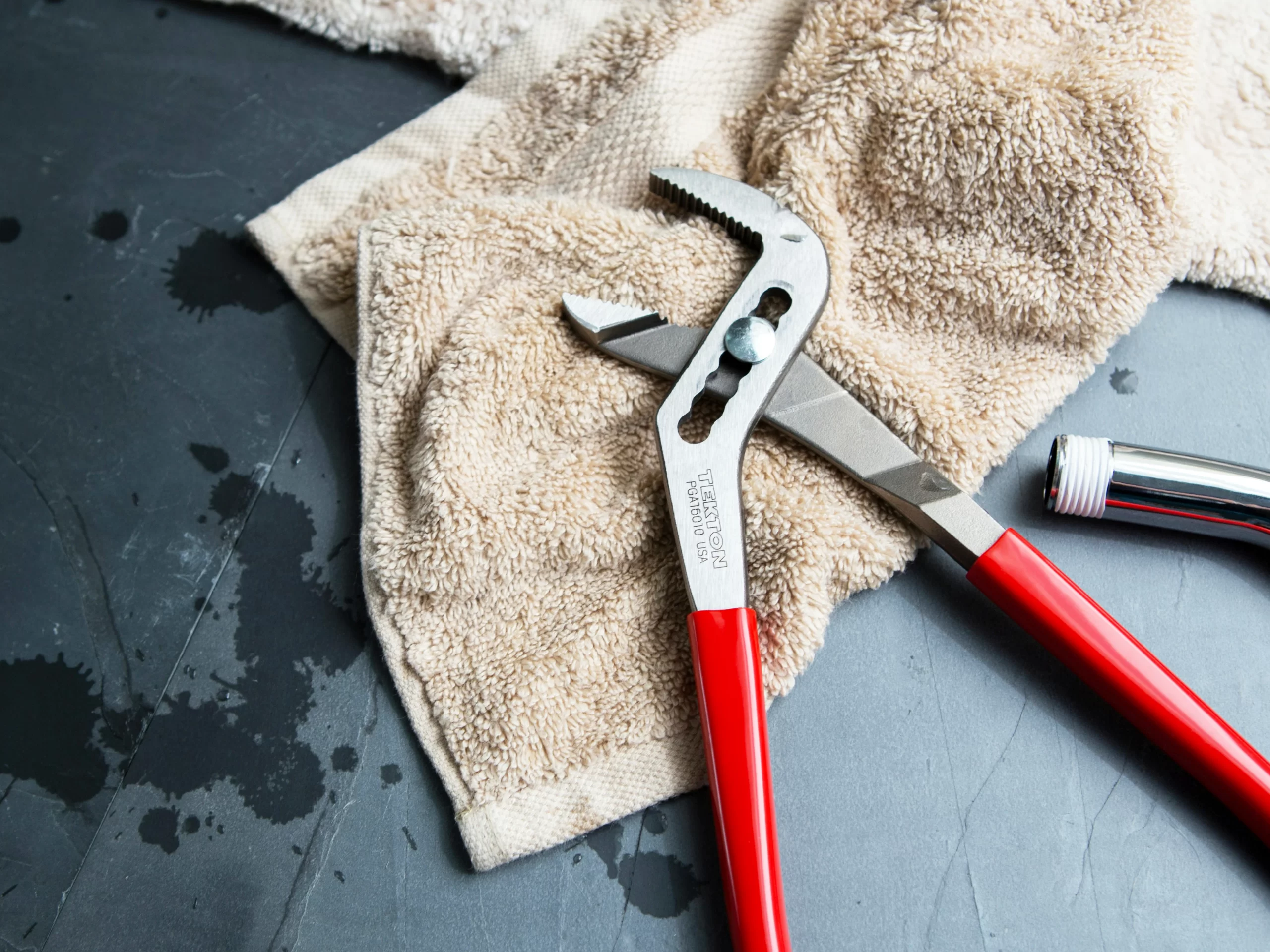 A pair of red-handled pliers and a metal pipe fitting rest on a beige towel, spread across a dark, wet surface with visible water drops—suggesting an emergency plumber is needed for urgent repairs.