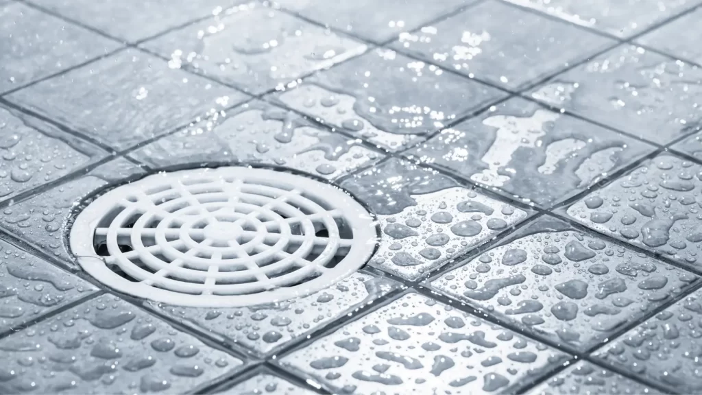 A close-up of a wet tiled floor with water droplets and a white circular drain cover in the center. The tiles are light-colored and the scene suggests a recently used shower or wet area.
