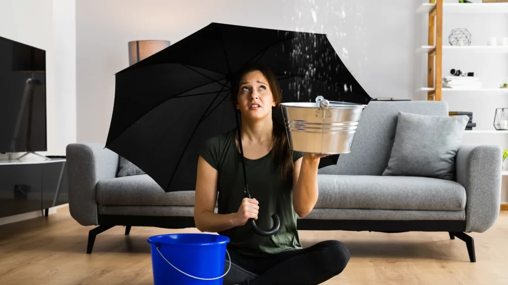 A woman sitting on the floor in a living room holds an umbrella and a metal bucket to catch water leaking from the ceiling, looking up worriedly. A blue bucket is placed near her on the floor.