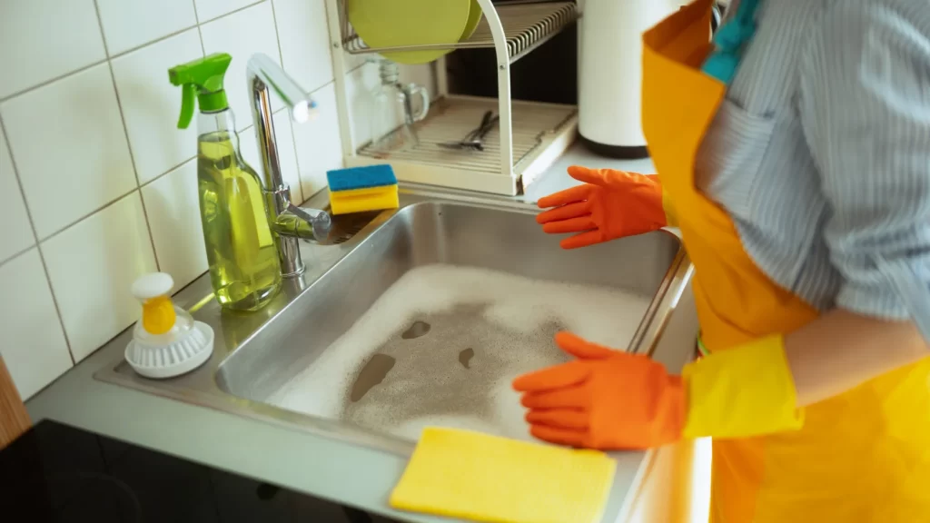 Person wearing orange gloves and a yellow apron stands by a kitchen sink filled with soapy water; cleaning supplies, sponges, and a dish rack are visible on the counter.