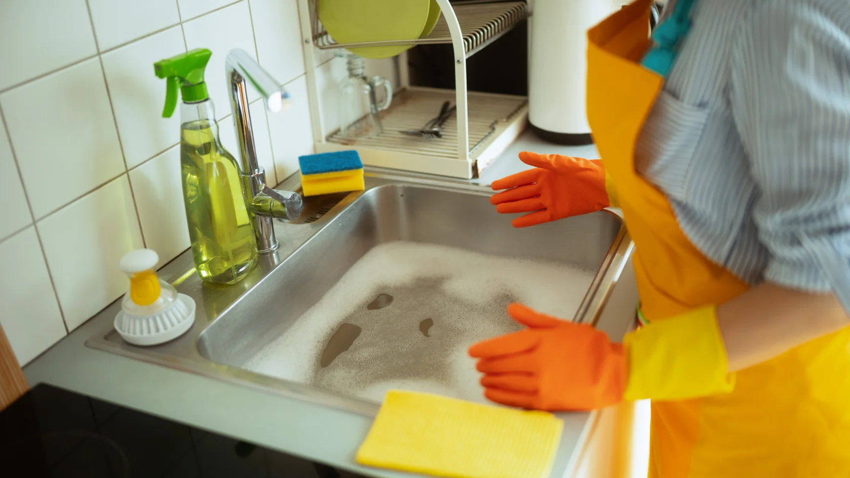 Person wearing orange gloves and a yellow apron stands by a kitchen sink filled with soapy water; cleaning supplies, sponges, and a dish rack are visible on the counter.