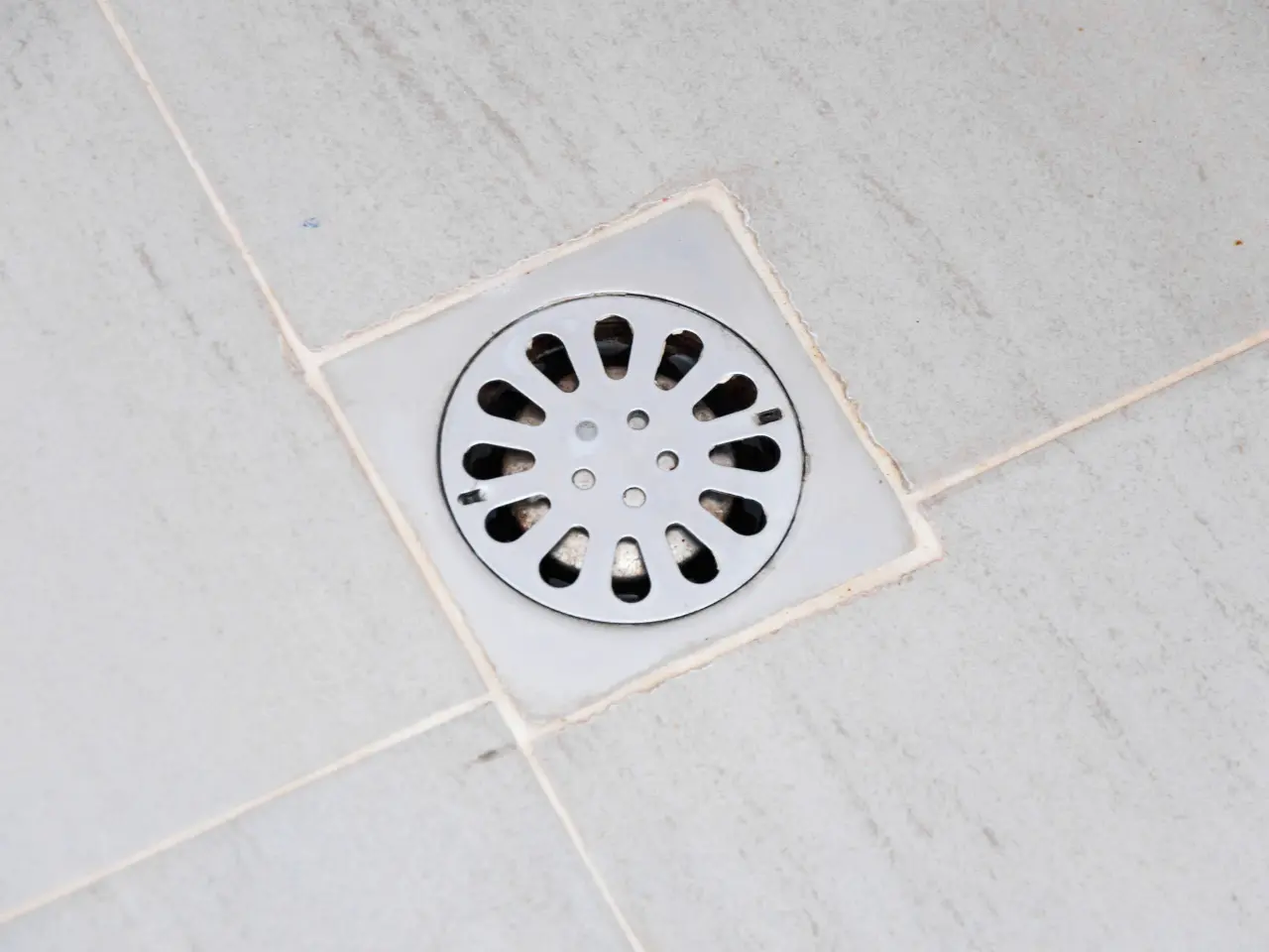 A close-up of a circular metal floor drain cover with oval-shaped holes, set in light gray tiled flooring. The cover is surrounded by a square tile border with visible grout lines.