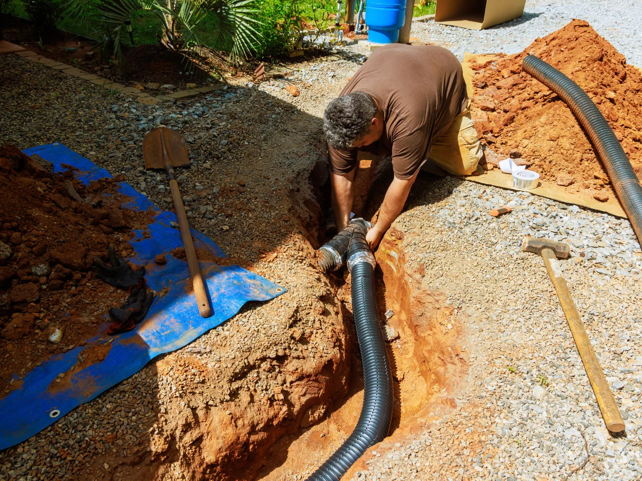A man installs a black drainage pipe in a trench surrounded by dirt and gravel. A shovel, hammer, gloves, and blue tarp are nearby. Sunlight illuminates the work area with plants in the background.