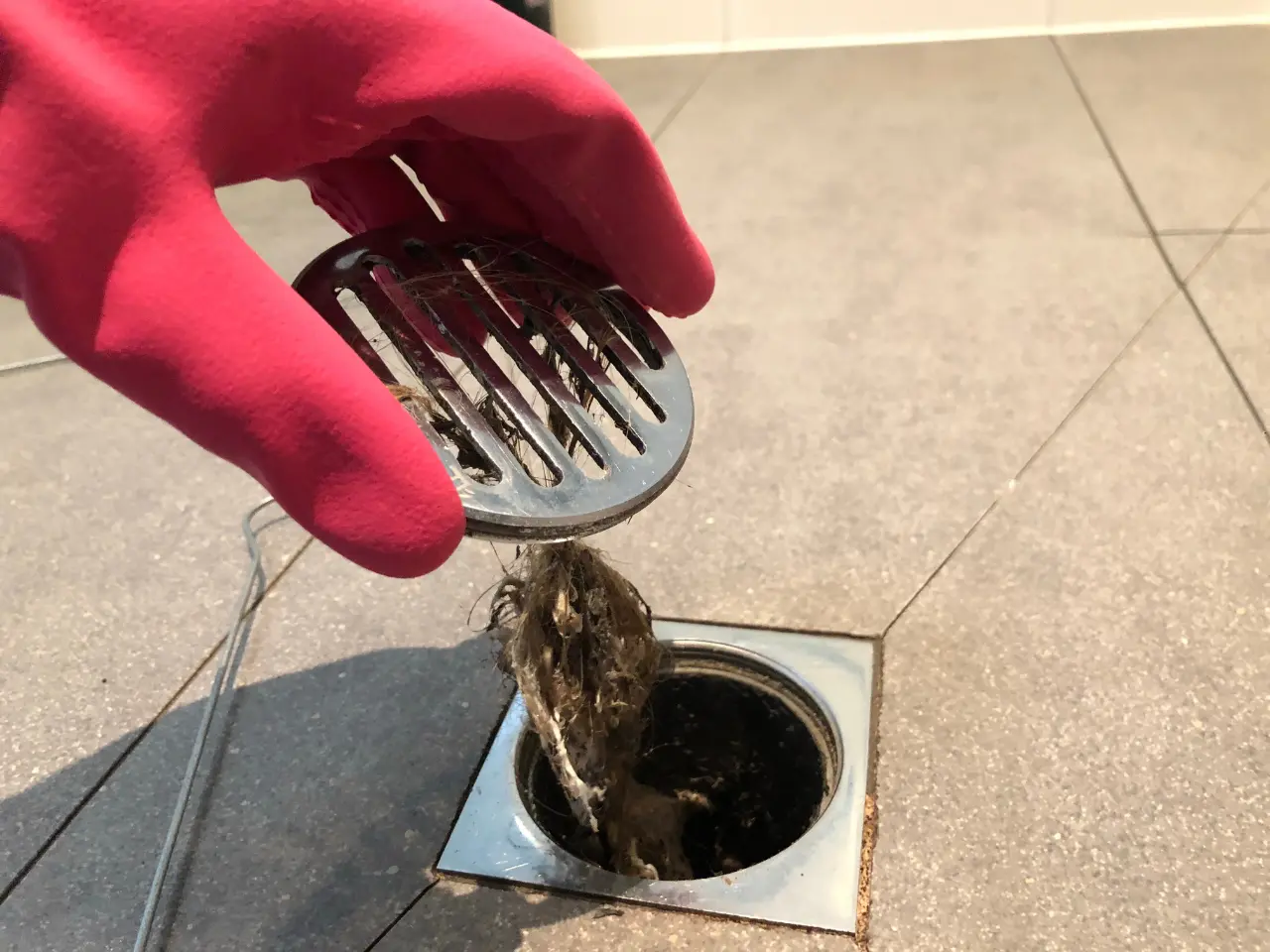 A hand wearing a pink rubber glove lifts a metal drain cover, revealing a clump of hair and debris caught in the bathroom floor drain.