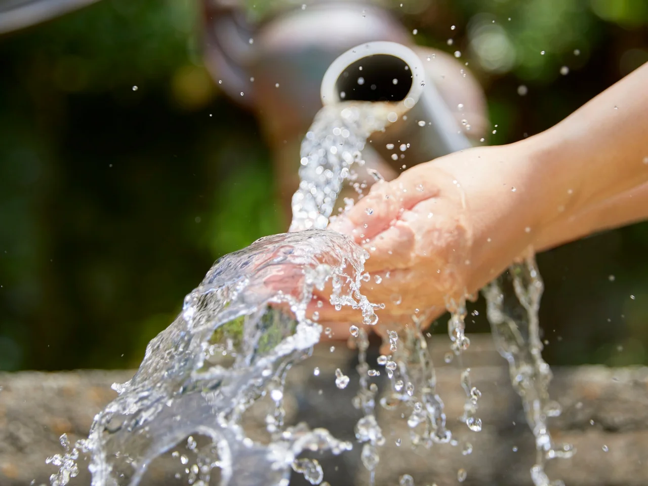 Close-up of hands being washed under a stream of flowing water, with droplets splashing around and a blurred green background.
