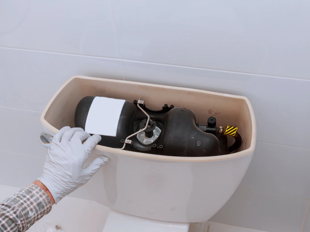 A person wearing a white glove lifts the lid of a toilet tank, revealing a black mechanism inside the tank against a white tiled wall.