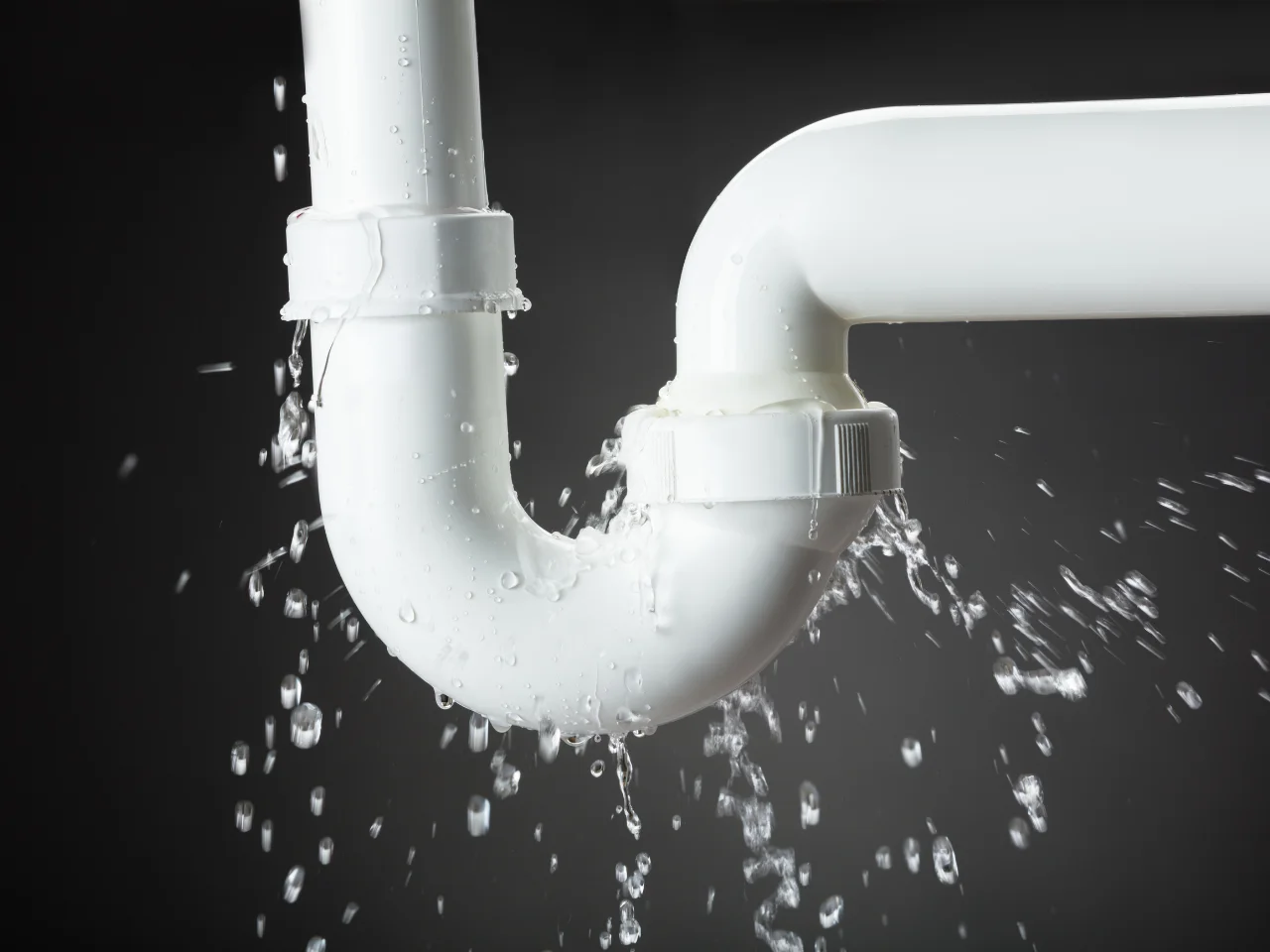 White plastic pipe with a U-bend is leaking water at a joint, causing water to spray out and drip, set against a dark background.