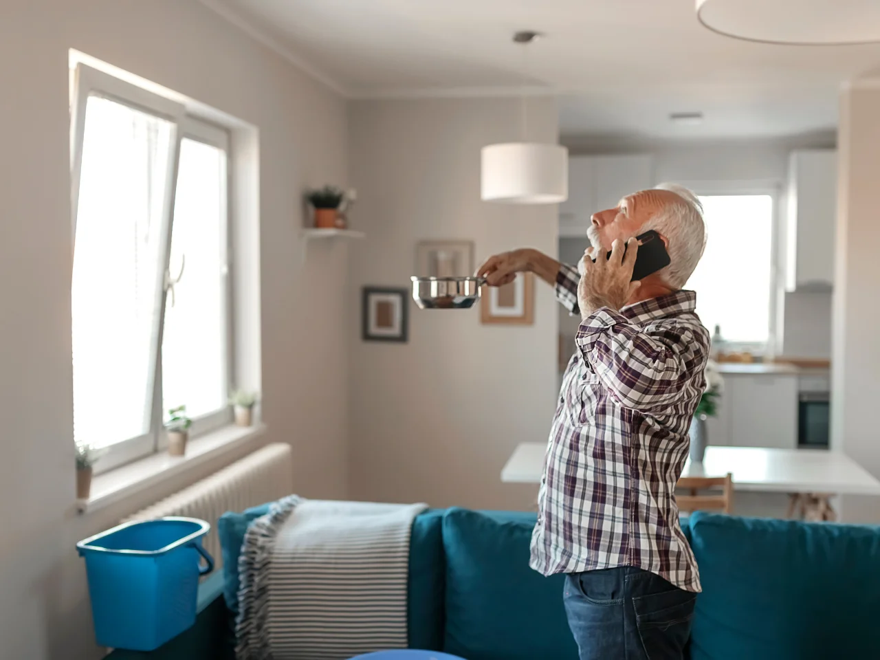 An older man stands in his living room, holding a pot under a leaking ceiling while talking on the phone. A blue bucket is on the couch, and the room appears bright with modern decor.