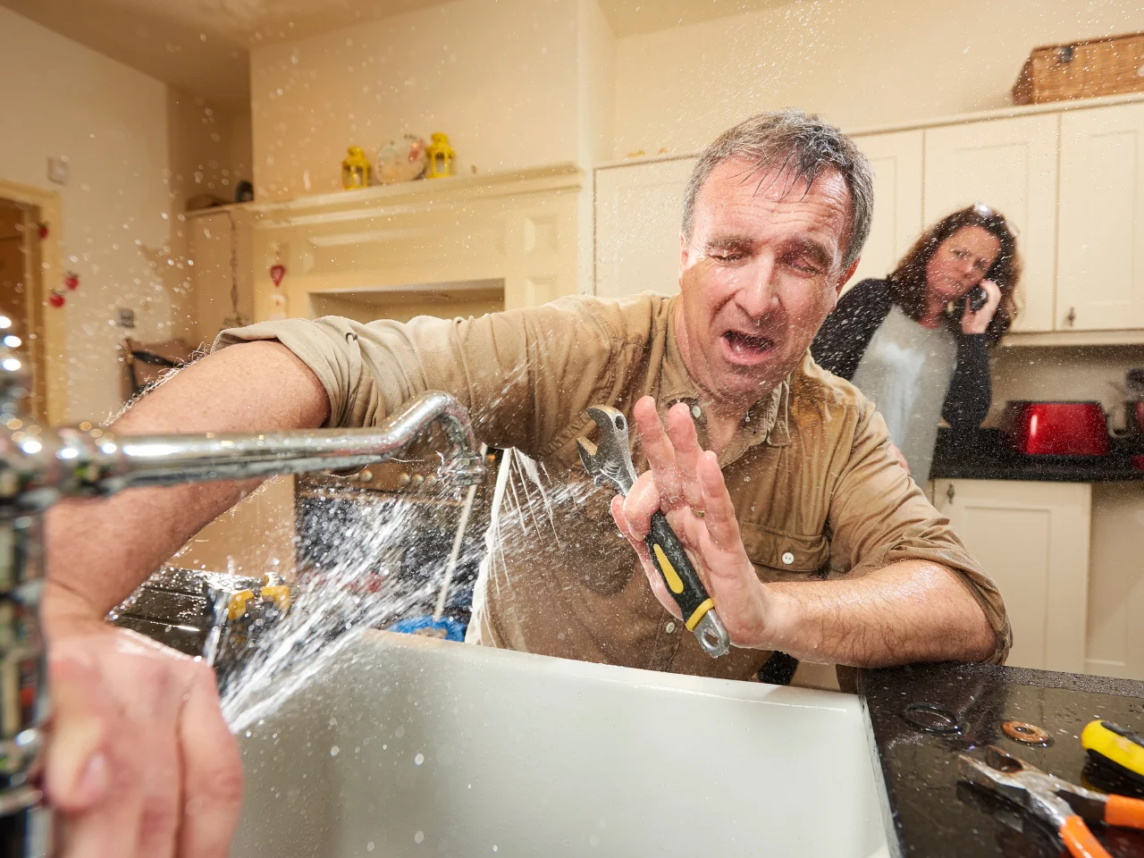 A man struggles to fix a kitchen sink as water sprays everywhere, soaking him. He looks frustrated and holds a wrench. In the background, a woman is on the phone, appearing concerned.