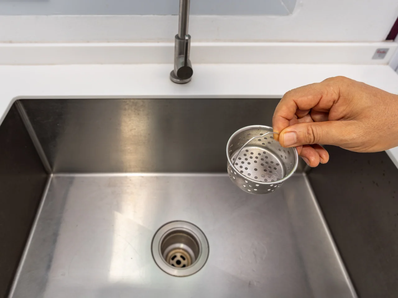 A hand holds a round metal sink strainer above a stainless steel kitchen sink, with the sink drain visible at the bottom.