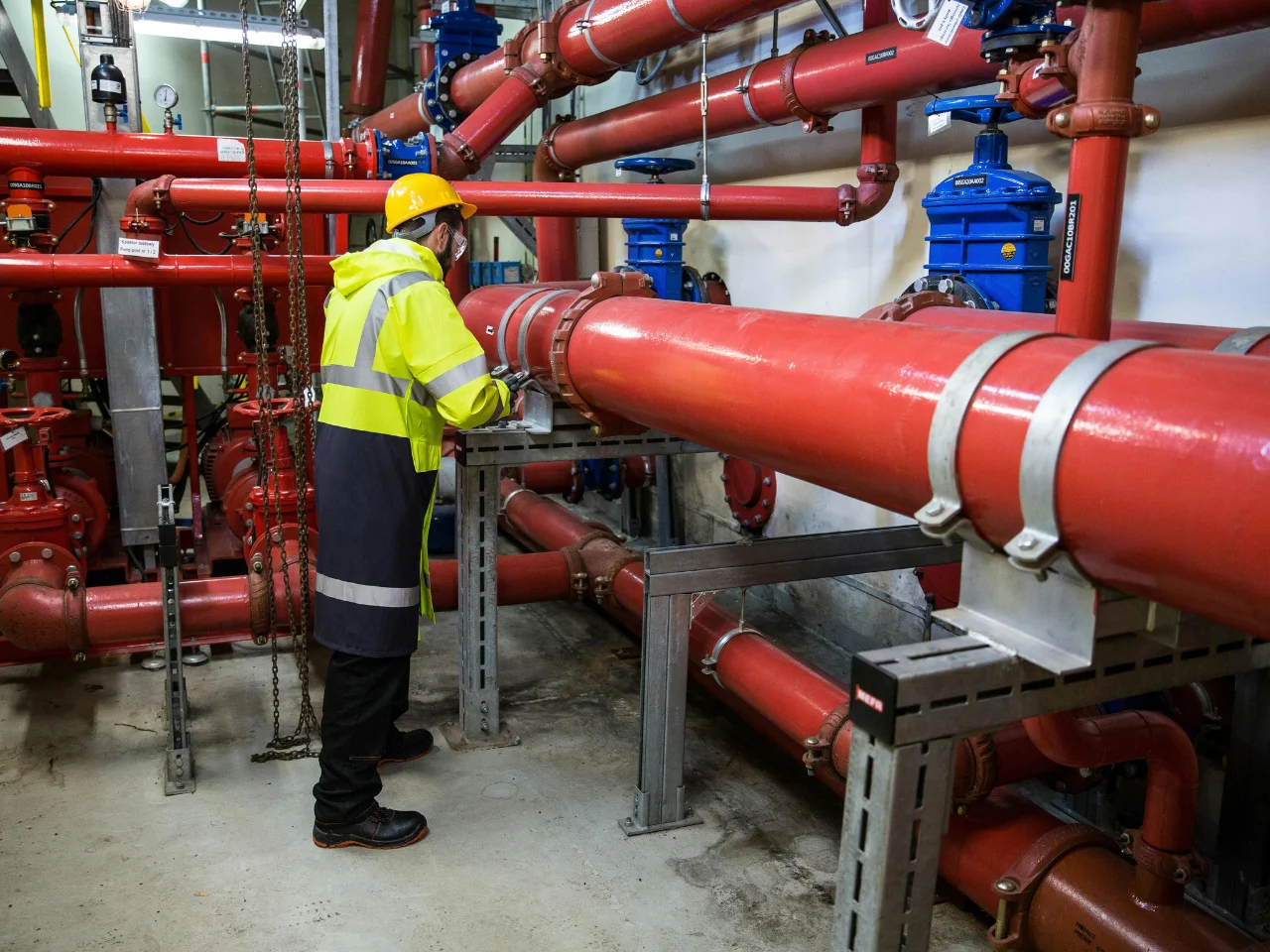 A worker in a yellow safety jacket and helmet inspects large red industrial pipes and valves in a mechanical room.