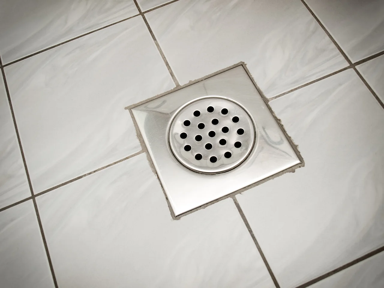 A close-up of a square metal floor drain with circular holes, set in white ceramic tiles with gray veining. The grout lines form a grid pattern around the drain.