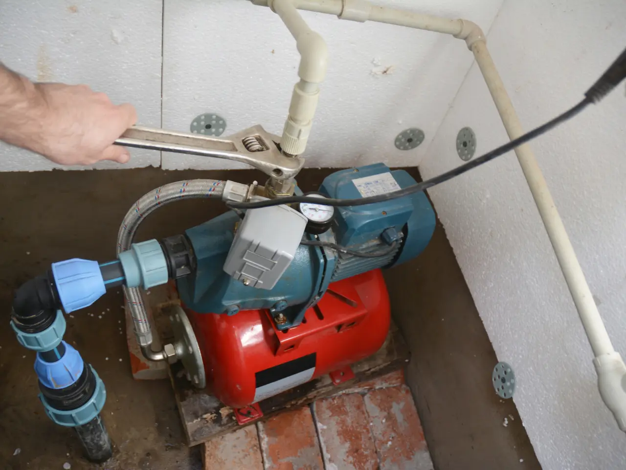 A person uses a wrench to adjust a valve on a blue and red water pump, which is connected to white PVC pipes in a small, tiled utility area.