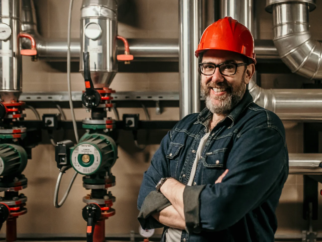 A man wearing a red hard hat, glasses, and a denim shirt stands with arms crossed, smiling in front of industrial metal pipes and valves inside a mechanical room.