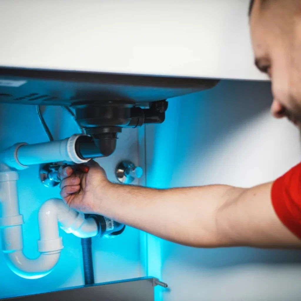 A person in a red shirt repairs white plumbing pipes under a sink, adjusting a connection with their hand. The workspace, illuminated by blue-tinted light, highlights expert drain repair and plumbing in Toronto.