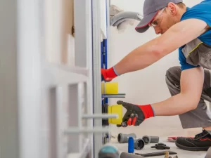 A plumber wearing gloves, safety glasses, and a cap installs or repairs plumbing pipes and fittings in a bathroom, with various tools scattered on the floor nearby—expertly handling drain repair and plumbing in Toronto.