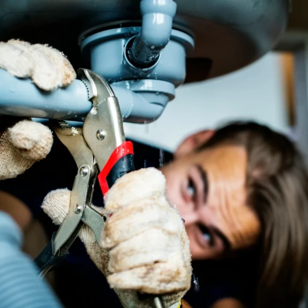 A person wearing gloves uses pliers to tighten or fix a pipe under a sink, focusing intently on plumbing work and drain repair in Toronto.