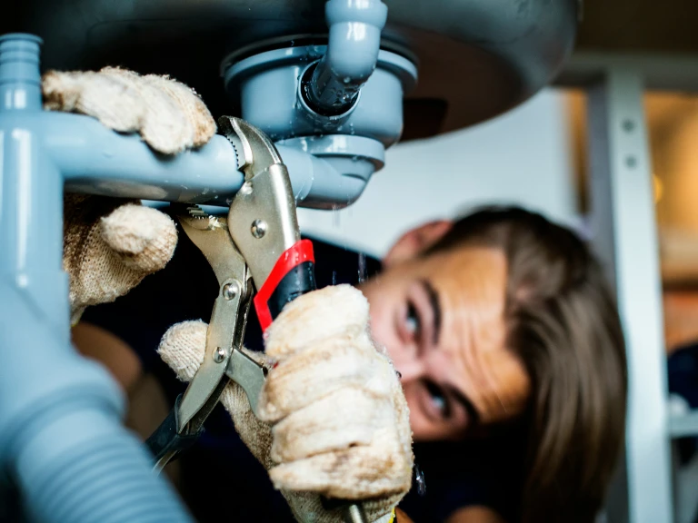 A person wearing gloves uses pliers to tighten or fix a pipe under a sink, focusing intently on plumbing work and drain repair in Toronto.