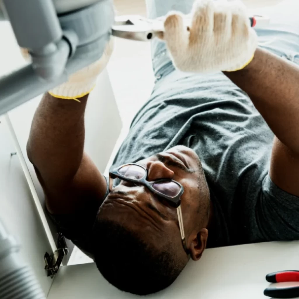 A man wearing glasses and gloves lies on his back under a sink, using pliers for drain repair and plumbing in Toronto. Tools are visible on the floor beside him.