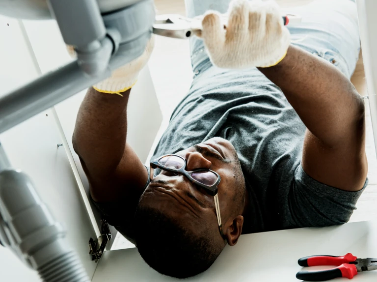 A man wearing glasses and gloves lies on his back under a sink, using pliers for drain repair and plumbing in Toronto. Tools are visible on the floor beside him.