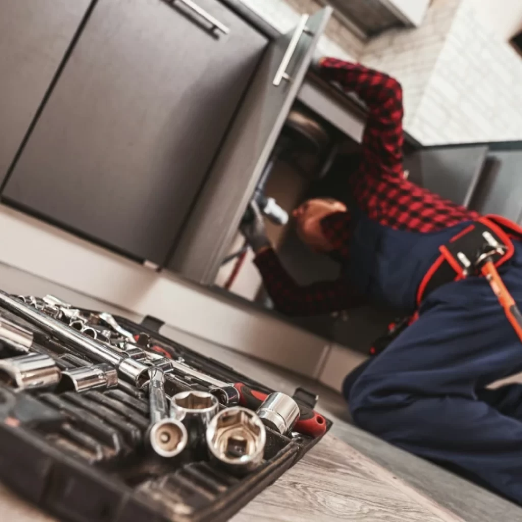 A plumber in a red plaid shirt and blue overalls is kneeling on the floor, working under a kitchen sink. A toolkit with various socket wrenches is open in the foreground, highlighting expert drain repair and plumbing in Toronto.