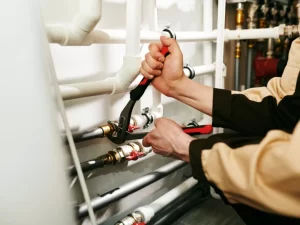 A plumber in Mississauga, wearing a beige and brown uniform, uses pliers and a wrench to tighten plumbing pipes and fittings indoors, focusing on maintenance or repair work.