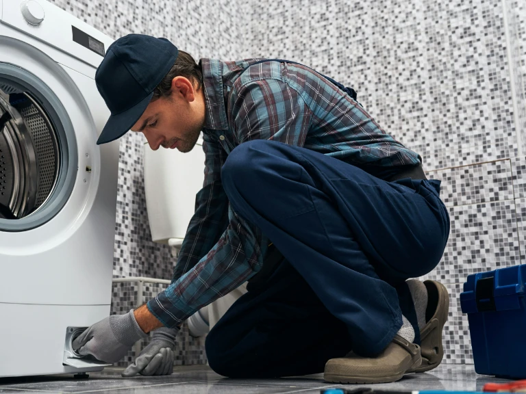 A repairman in a plaid shirt, blue pants, and a cap kneels on a bathroom floor, fixing a washing machine. He uses tools from a blue toolbox beside him, handling drain repair as part of expert plumbing in Toronto. Tiled walls and a toilet are in the background.