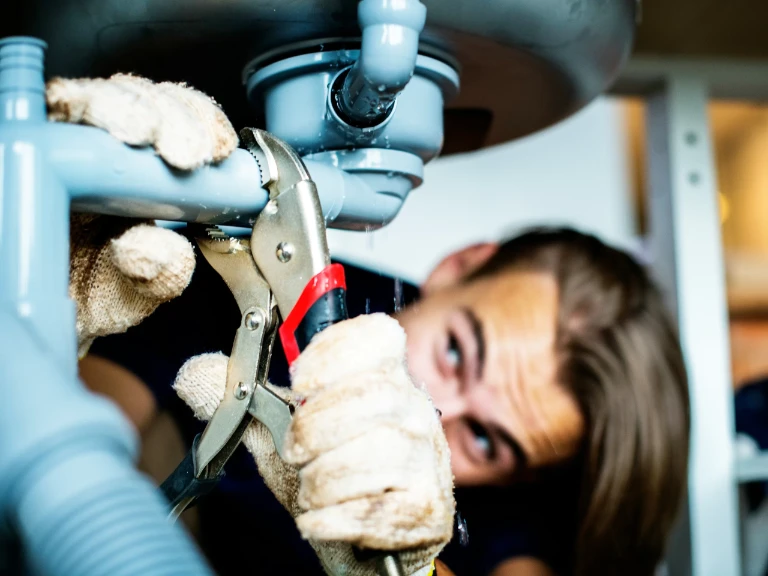 A plumber Toronto wears gloves and uses pliers to tighten a pipe under a sink, focusing closely on the plumbing work.