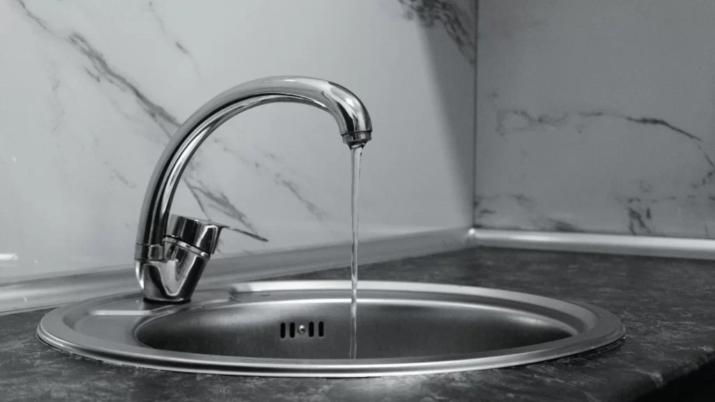 A stainless steel kitchen sink with a curved faucet, running a thin stream of water, set against a countertop and white marble-patterned backsplash.