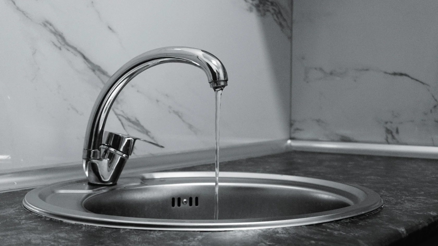A stainless steel kitchen sink with a curved faucet, running a thin stream of water, set against a countertop and white marble-patterned backsplash.
